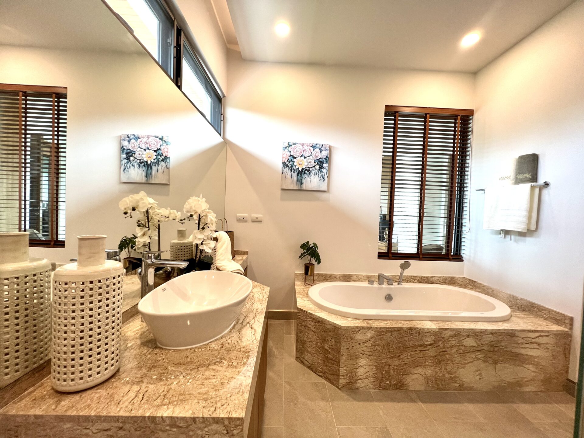 Spa-like bathroom with marble countertop, white vessel sink, woven baskets, orchids, and a built-in marble tub under two windows with wooden blinds.