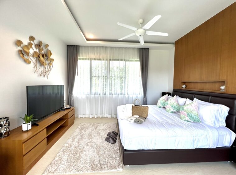 Bright hotel-style bedroom with a king bed, white linens and tropical-pattern pillows, a wooden TV console, and gold leaf wall decor by a large window with sheer curtains.