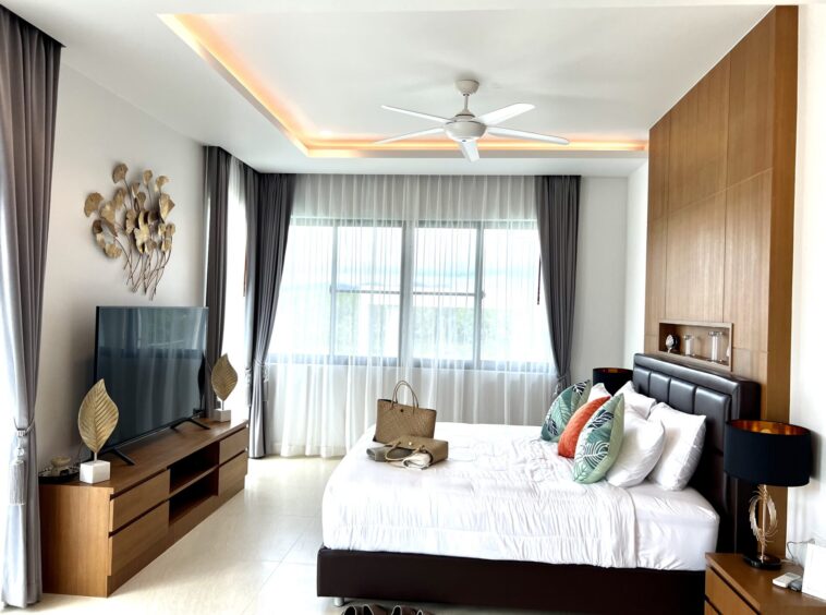 Modern hotel-style bedroom with a large bed, white bedding and colorful tropical cushions, a ceiling fan, and tall wooden wardrobes beside a window wall with sheer and dark curtains.