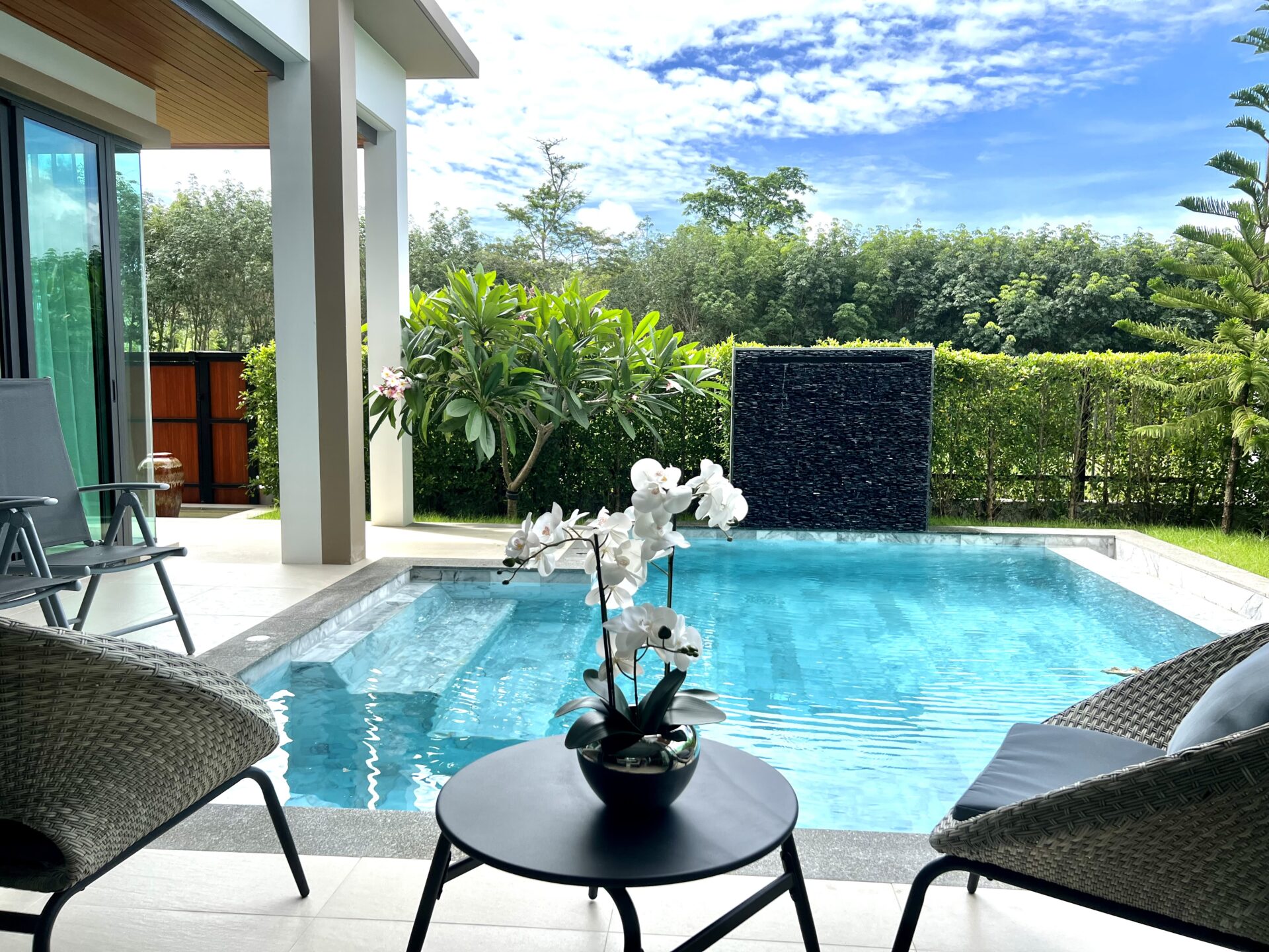 Poolside patio with wicker lounge chairs, a round table with an orchid centerpiece, and a bright blue pool water edge.