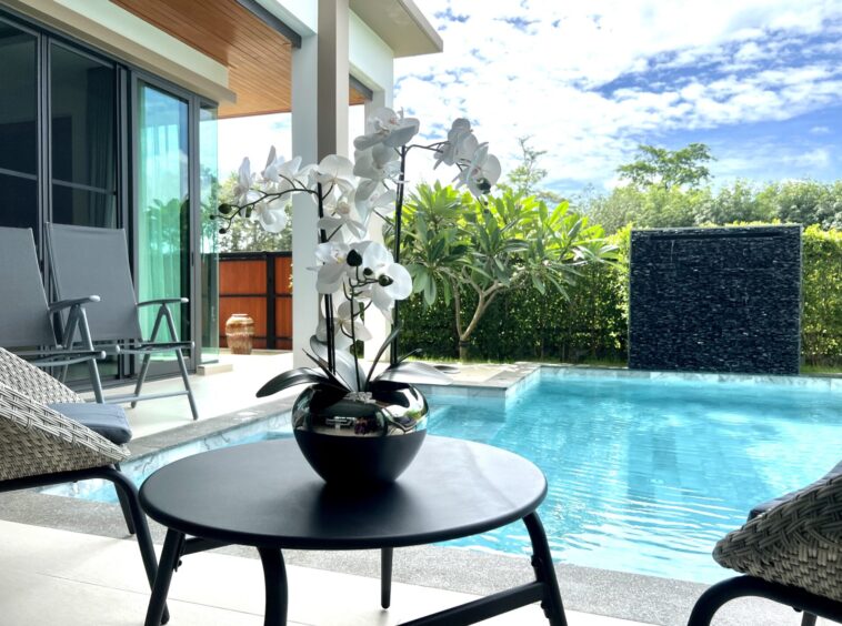 Poolside patio with a round black table holding a white orchid arrangement; lounge chairs nearby and sliding glass doors open to indoors.