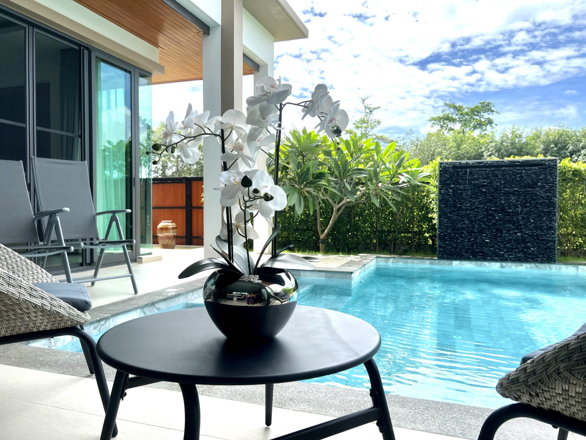 Poolside patio with a round black table holding a white orchid arrangement; lounge chairs nearby and sliding glass doors open to indoors.