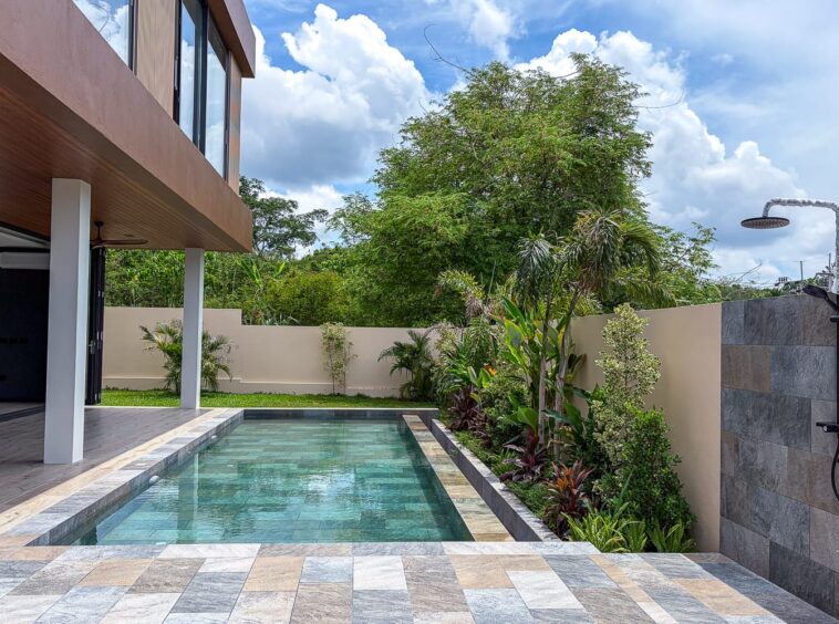 Modern backyard with a rectangular pool, stone-tiled deck, and lush greenery along a beige privacy wall.