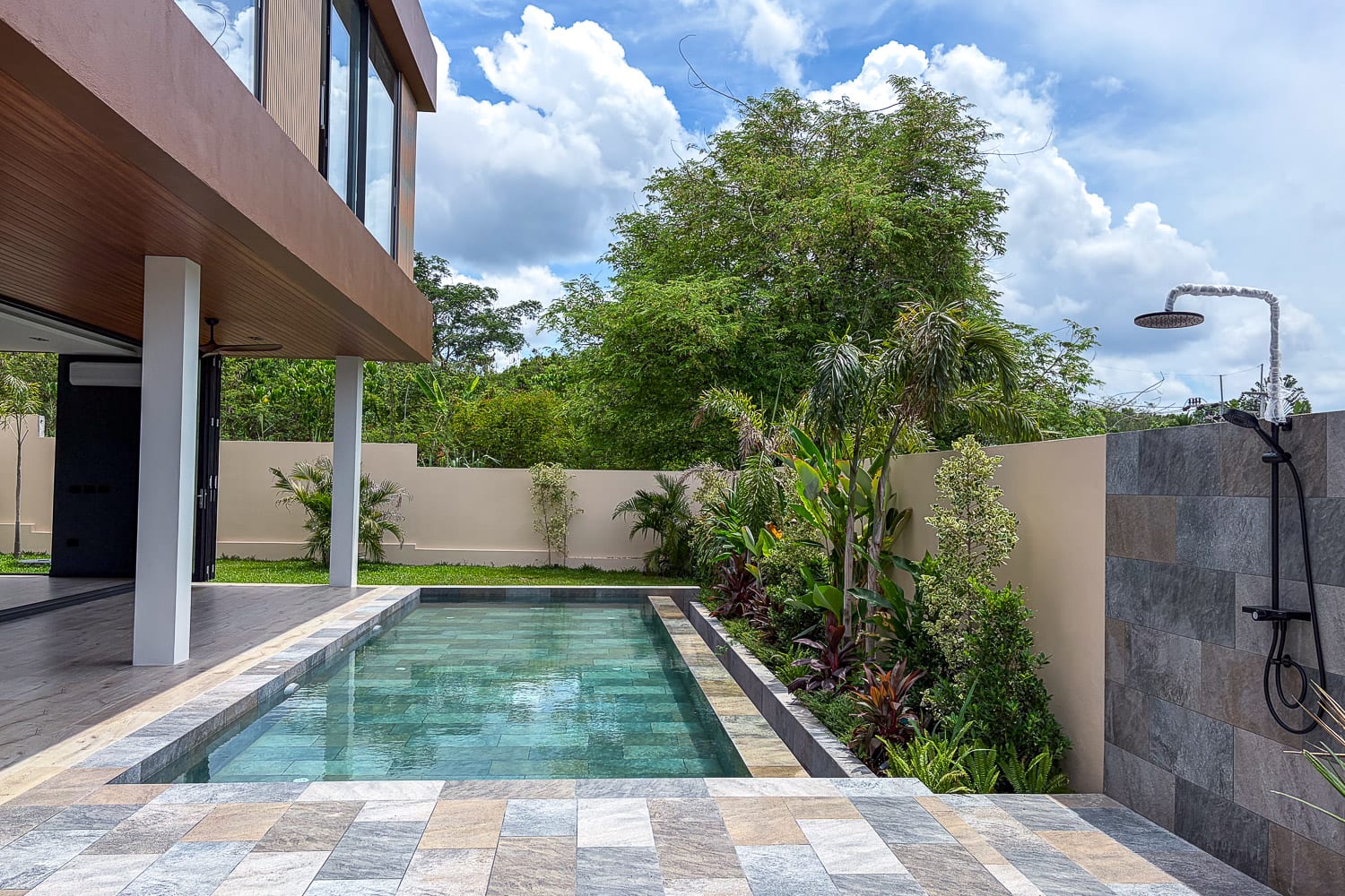 Modern backyard with a rectangular pool, stone-tiled deck, and lush greenery along a beige privacy wall.