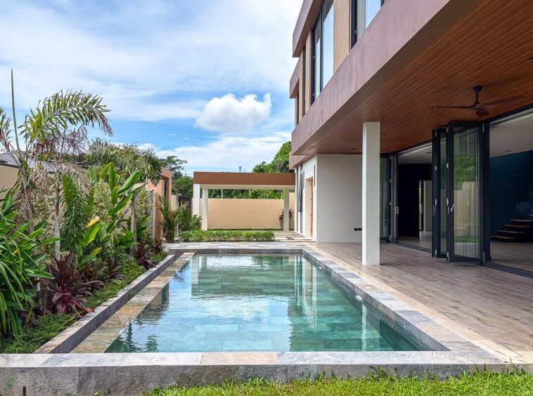 Rectangular pool beside a modern house with a wood-paneled ceiling, glass doors, and lush tropical plants along the left edge.