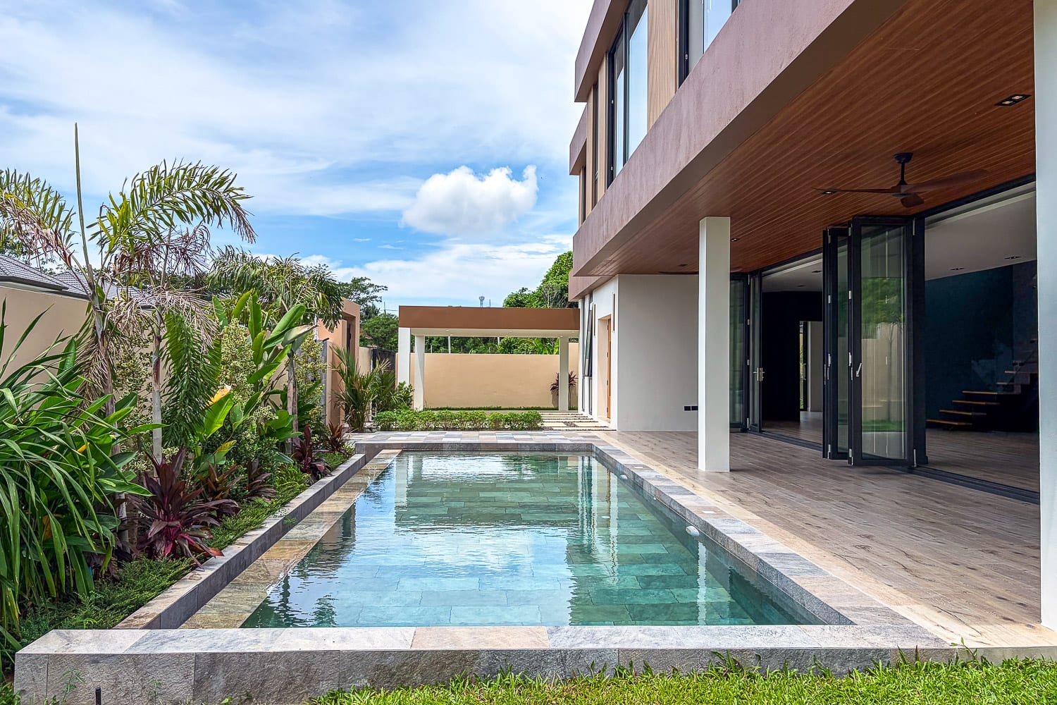 Rectangular pool beside a modern house with a wood-paneled ceiling, glass doors, and lush tropical plants along the left edge.