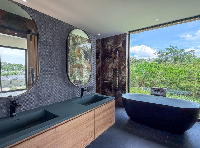 Modern bathroom with a double-sink vanity, dark green countertops, and a freestanding oval tub by a large window overlooking greenery.