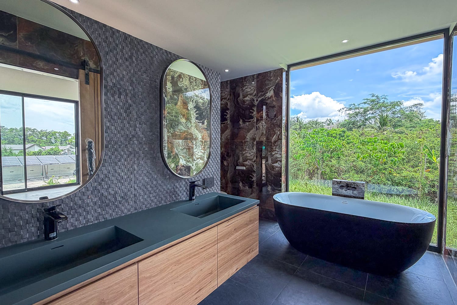 Modern bathroom with a double-sink vanity, dark green countertops, and a freestanding oval tub by a large window overlooking greenery.