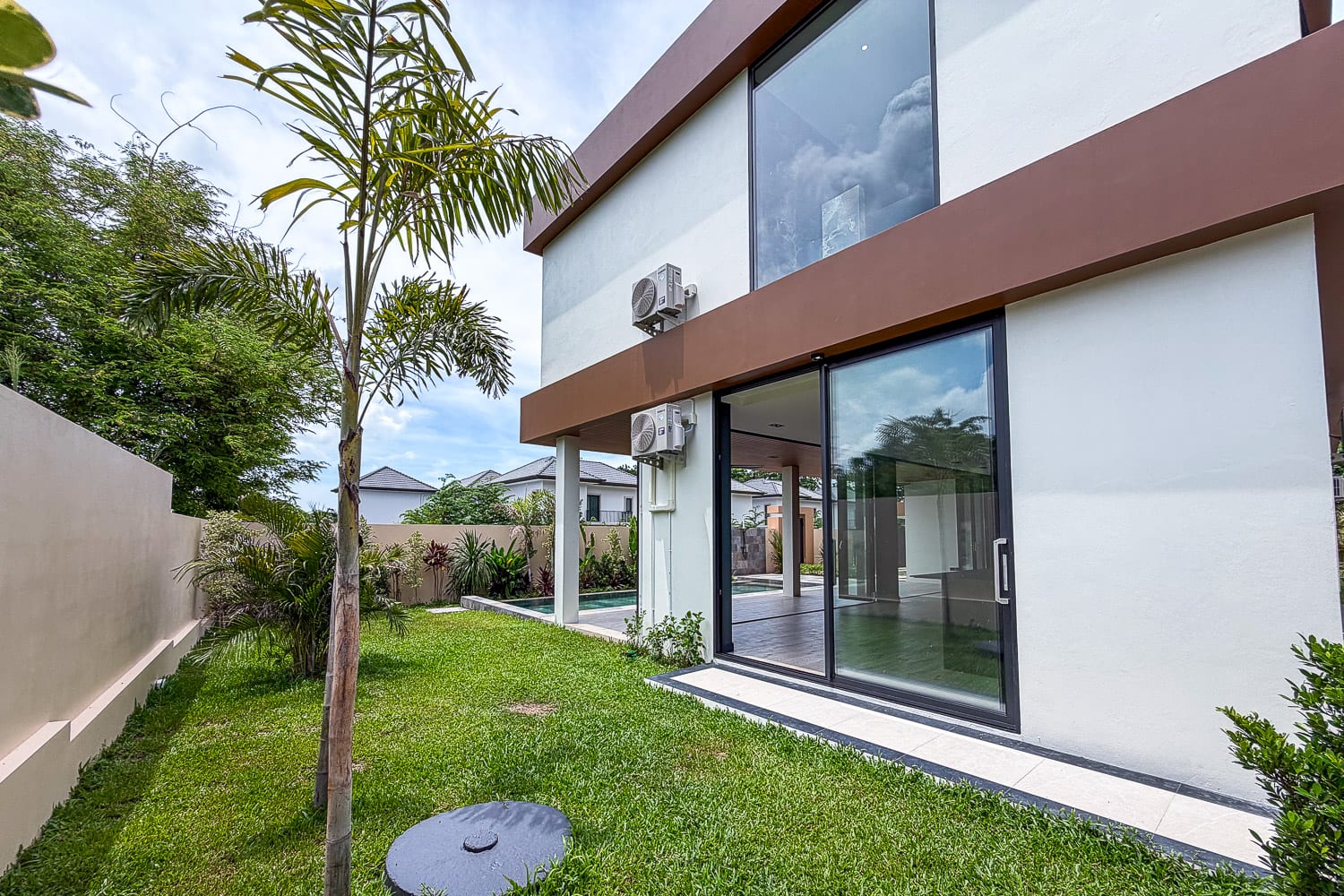 Modern two-story house with large glass sliding doors leading to a green backyard, a tall palm tree in the lawn, and exterior AC units mounted on the wall.