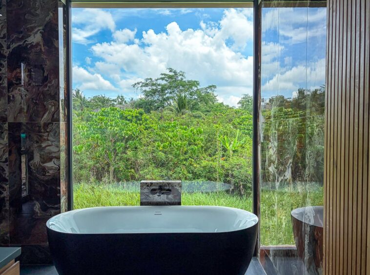 Freestanding black bathtub facing a large window with a lush green garden and blue sky outside, marble walls on the left and wooden paneling on the right.
