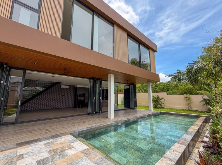 Modern two-story home with large glass doors opening to a rectangular pool and wooden deck. The house features a tan facade and a covered patio area.
