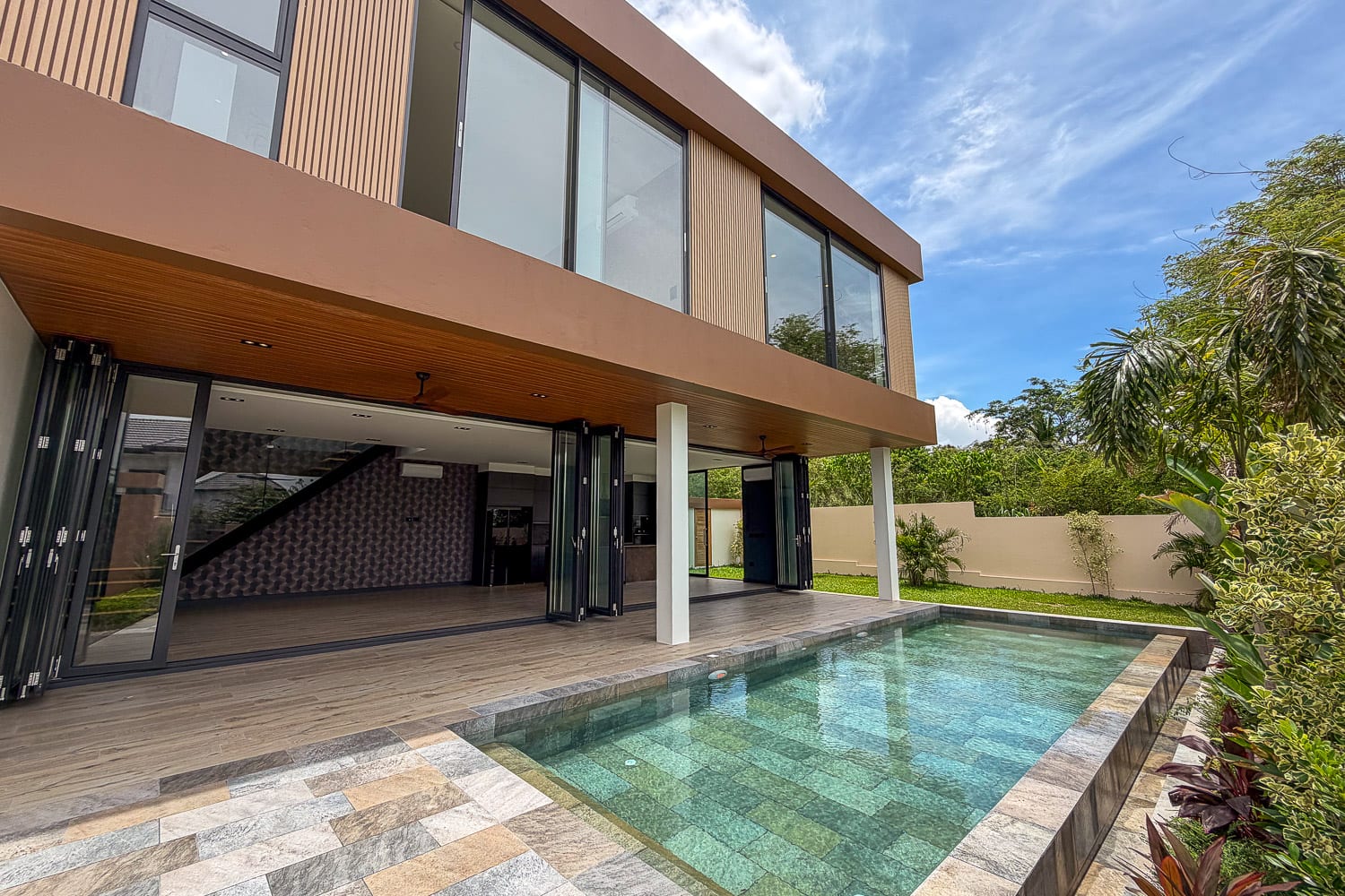 Modern two-story home with large glass doors opening to a rectangular pool and wooden deck. The house features a tan facade and a covered patio area.