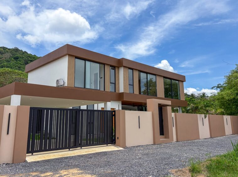Modern two-story house with a brown and beige exterior, large glass windows, and a black vertical-bar gate at a gravel driveway, set against green hills and a blue sky.