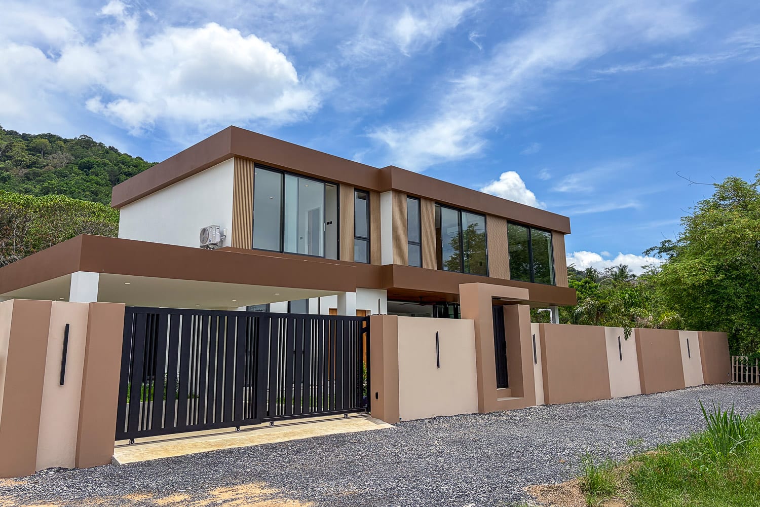 Modern two-story house with a brown and beige exterior, large glass windows, and a black vertical-bar gate at a gravel driveway, set against green hills and a blue sky.