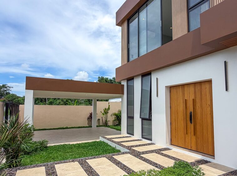 Modern two-story home with white walls, large glass windows, and a tall wooden front door under a brown overhang.