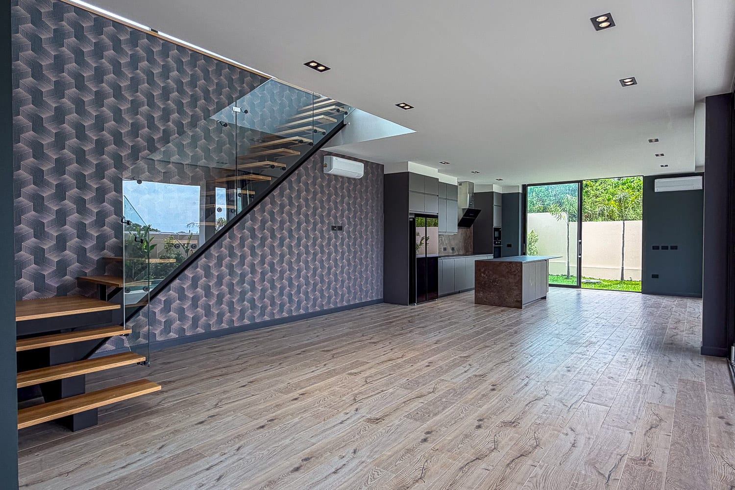 Open-plan living area with a glass-railed staircase, geometric-pattern wall, and a modern kitchen with island.