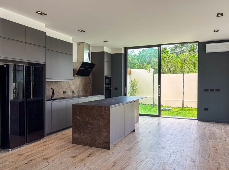 Modern kitchen with grey flat-panel cabinets, a dark island with stone countertop, stainless steel fridge, and a large glass door opening to a green backyard.