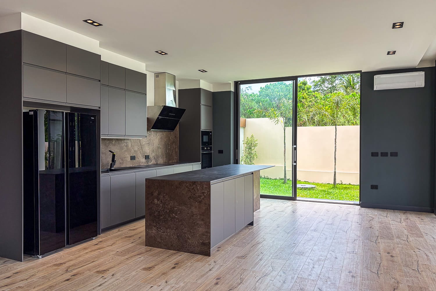 Modern kitchen with grey flat-panel cabinets, a dark island with stone countertop, stainless steel fridge, and a large glass door opening to a green backyard.