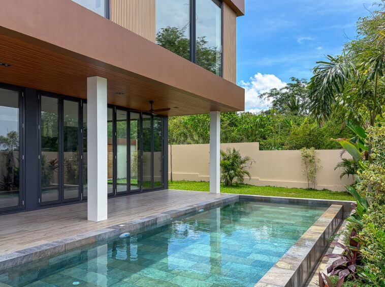 Modern house with a glass patio door facing a rectangular in-ground pool and wooden deck, surrounded by greenery under a blue sky.