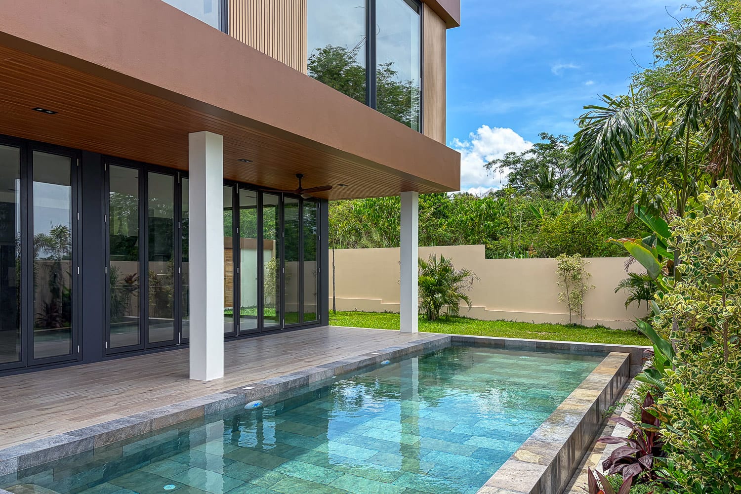 Modern house with a glass patio door facing a rectangular in-ground pool and wooden deck, surrounded by greenery under a blue sky.