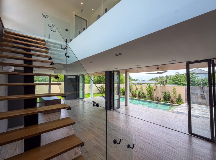 Modern open-plan interior with a floating wooden staircase and glass railing leading to an upper level, overlooking a bright living area and outdoor pool.