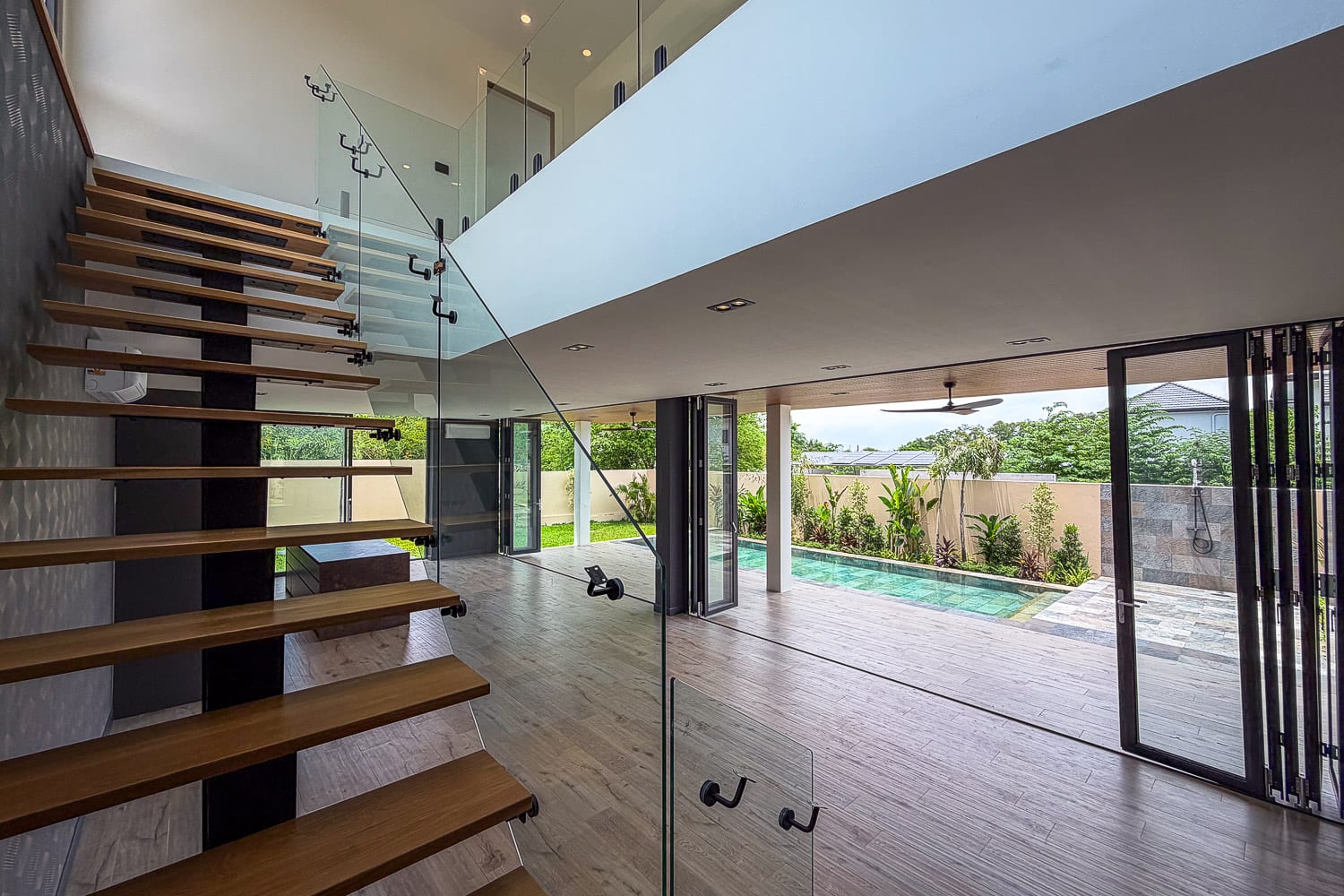 Modern open-plan interior with a floating wooden staircase and glass railing leading to an upper level, overlooking a bright living area and outdoor pool.
