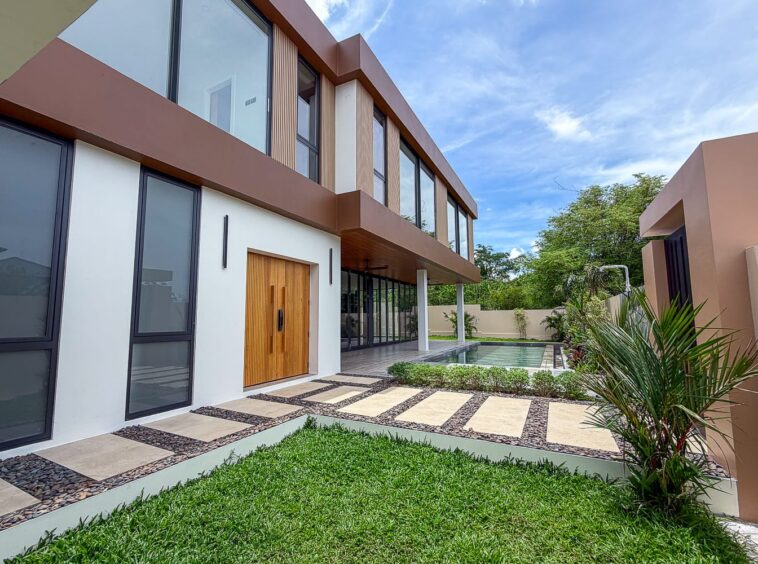 Modern two-story house with large glass windows, a wooden front door, and a rectangular pool along the back yard.