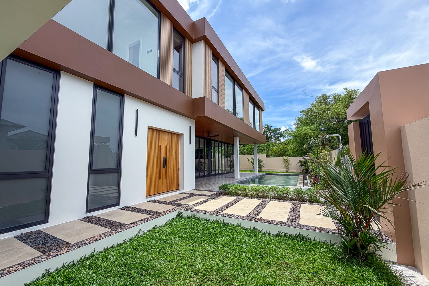 Modern two-story house with large glass windows, a wooden front door, and a rectangular pool along the back yard.