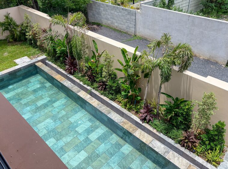 Rectangular turquoise-tiled pool with stone coping, surrounded by tropical plants and a beige privacy wall.