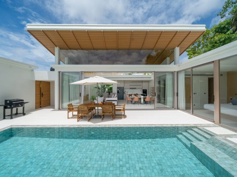 Modern backyard with a turquoise tiled pool, a wooden dining table under a white umbrella, and glass sliding doors opening to an indoor living/kitchen area.