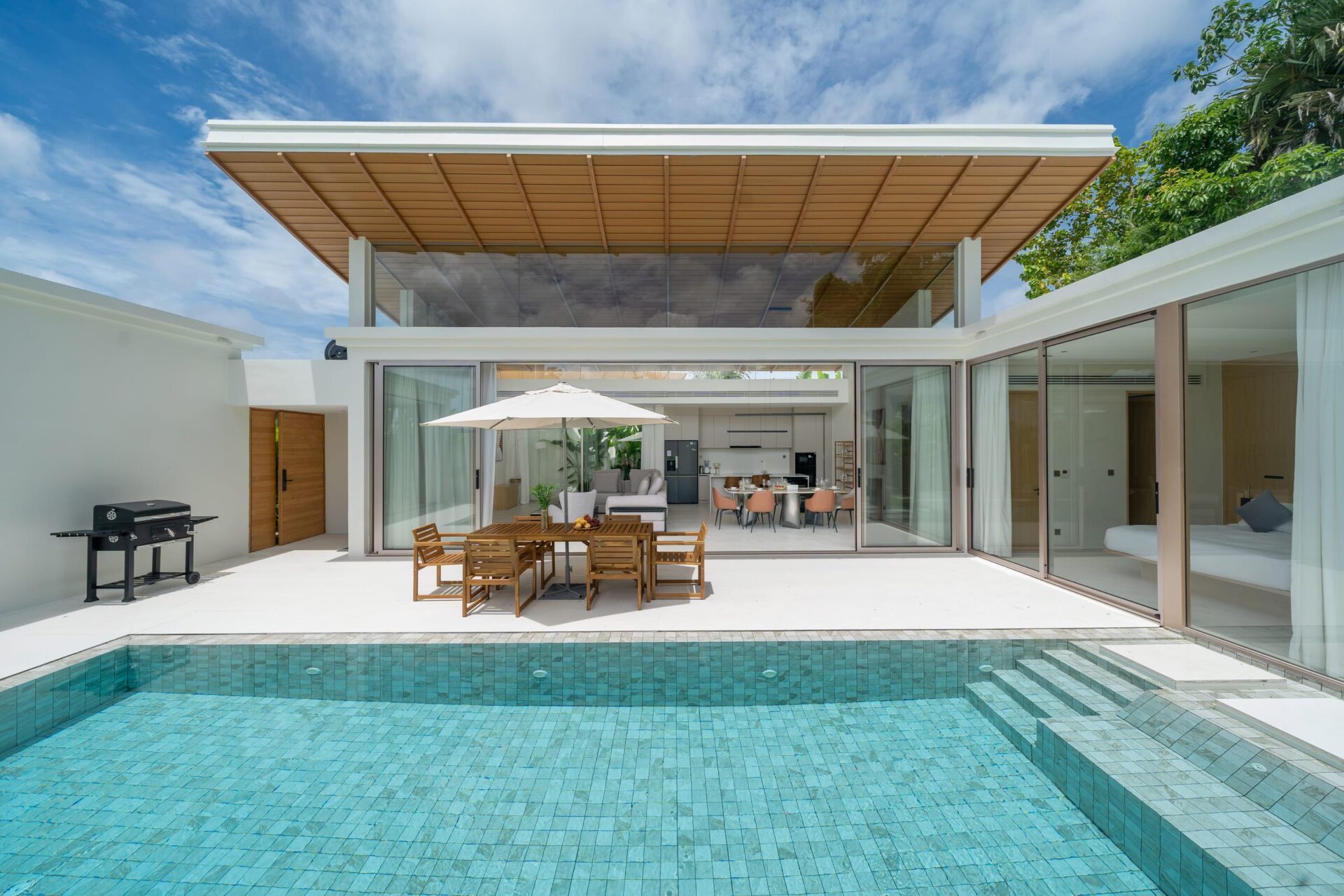 Modern backyard with a turquoise tiled pool, a wooden dining table under a white umbrella, and glass sliding doors opening to an indoor living/kitchen area.