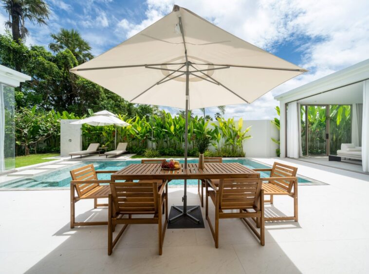 Outdoor patio with a wooden dining table and chairs under a large white umbrella beside a rectangular pool, surrounded by tropical plants.