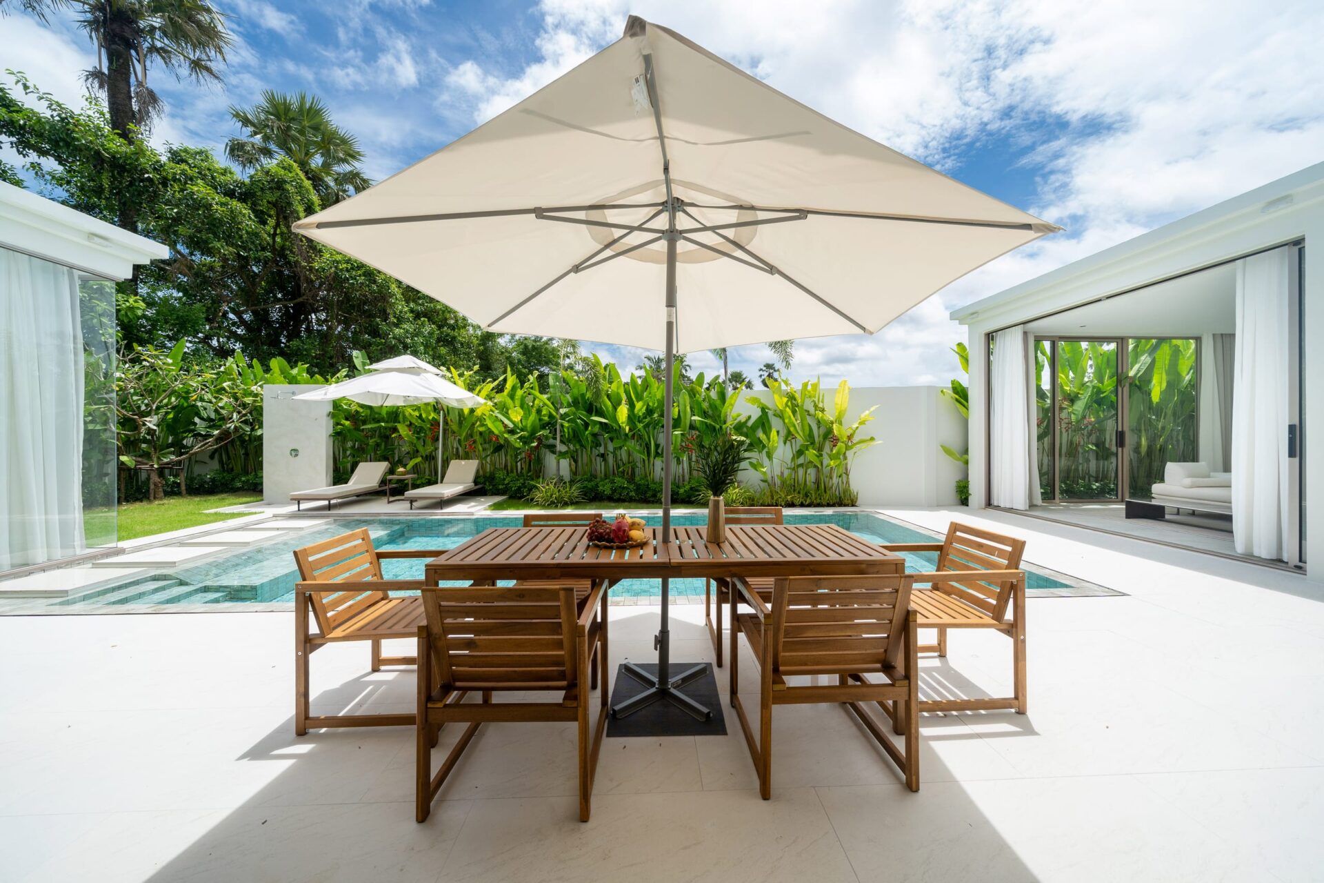 Outdoor patio with a wooden dining table and chairs under a large white umbrella beside a rectangular pool, surrounded by tropical plants.