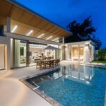 Modern house with an illuminated wooden pergola over a glass-walled living area, outdoor dining by a blue-tiled pool at dusk