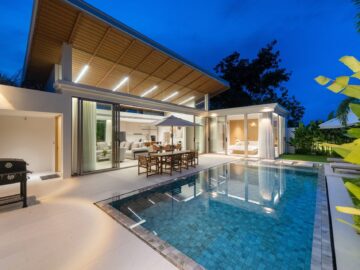 Modern house with an illuminated wooden pergola over a glass-walled living area, outdoor dining by a blue-tiled pool at dusk
