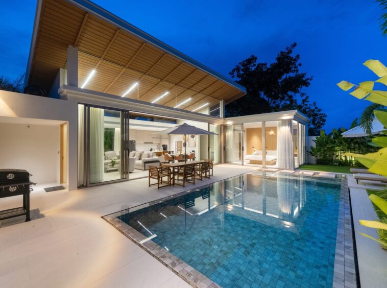 Modern house with an illuminated wooden pergola over a glass-walled living area, outdoor dining by a blue-tiled pool at dusk