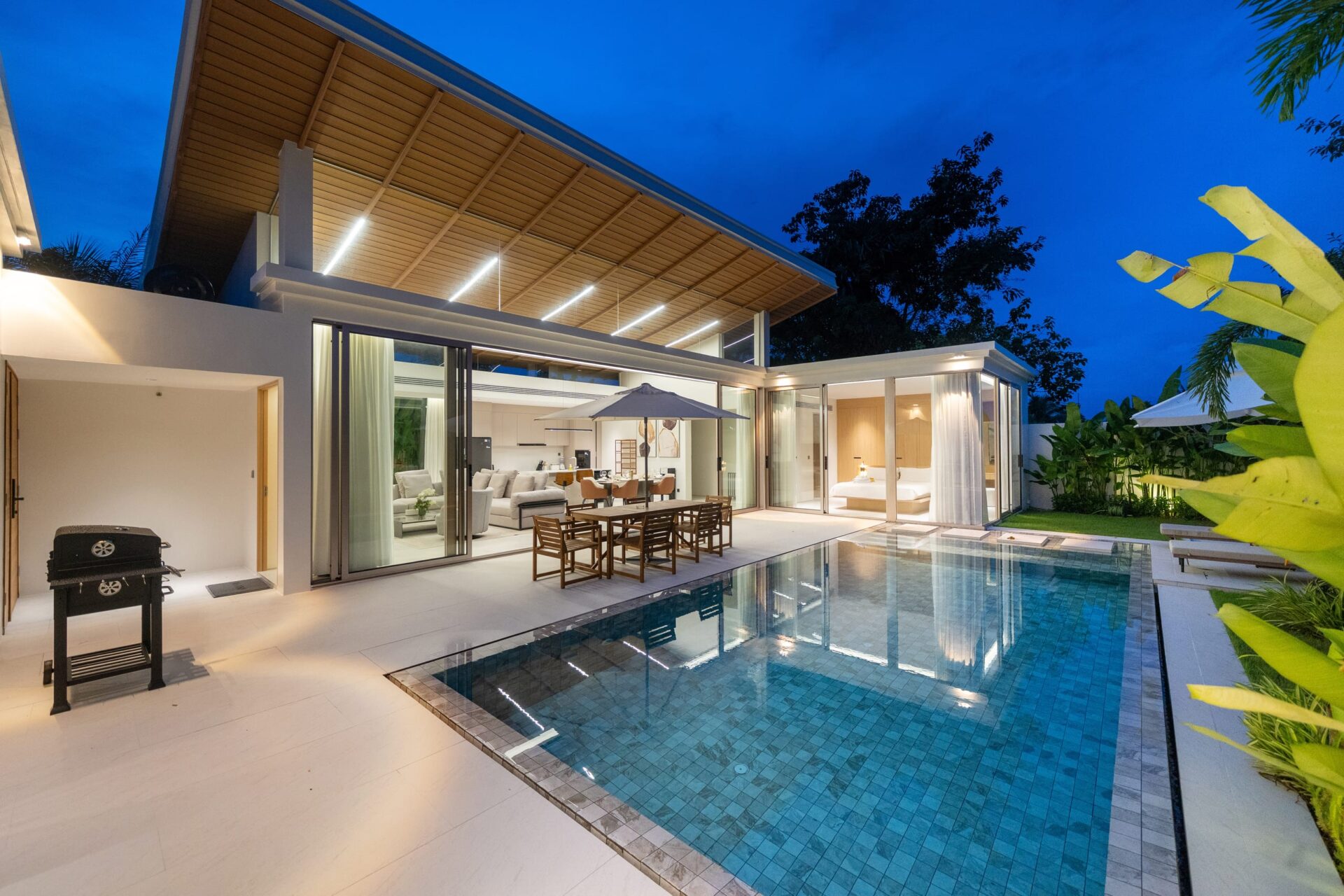 Modern house with an illuminated wooden pergola over a glass-walled living area, outdoor dining by a blue-tiled pool at dusk