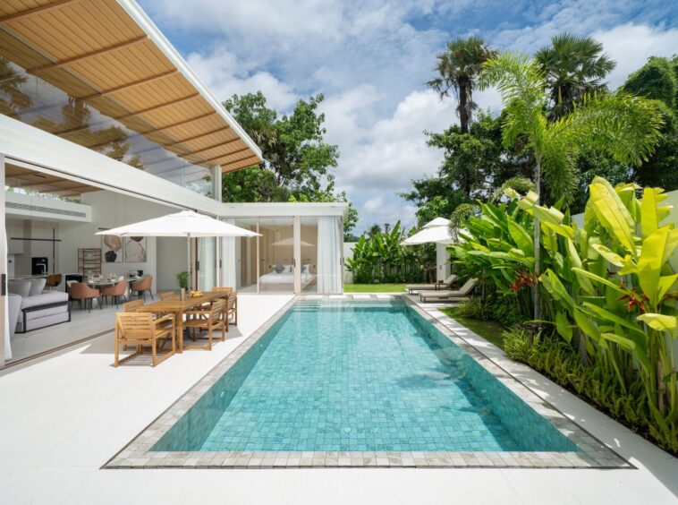 Modern backyard with a rectangular turquoise pool, glass-walled living area, wooden dining table under umbrellas, and tropical plants.