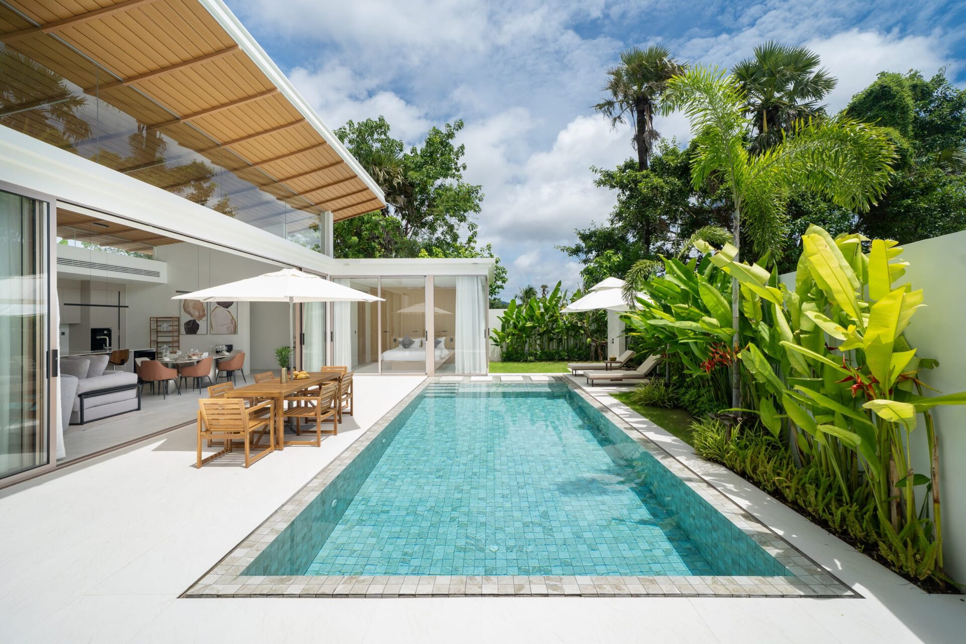 Modern backyard with a rectangular turquoise pool, glass-walled living area, wooden dining table under umbrellas, and tropical plants.