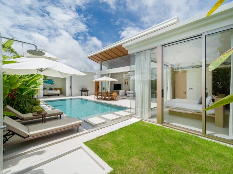 Modern villa backyard with a rectangular pool, white loungers, and umbrellas beside a glass-walled living area