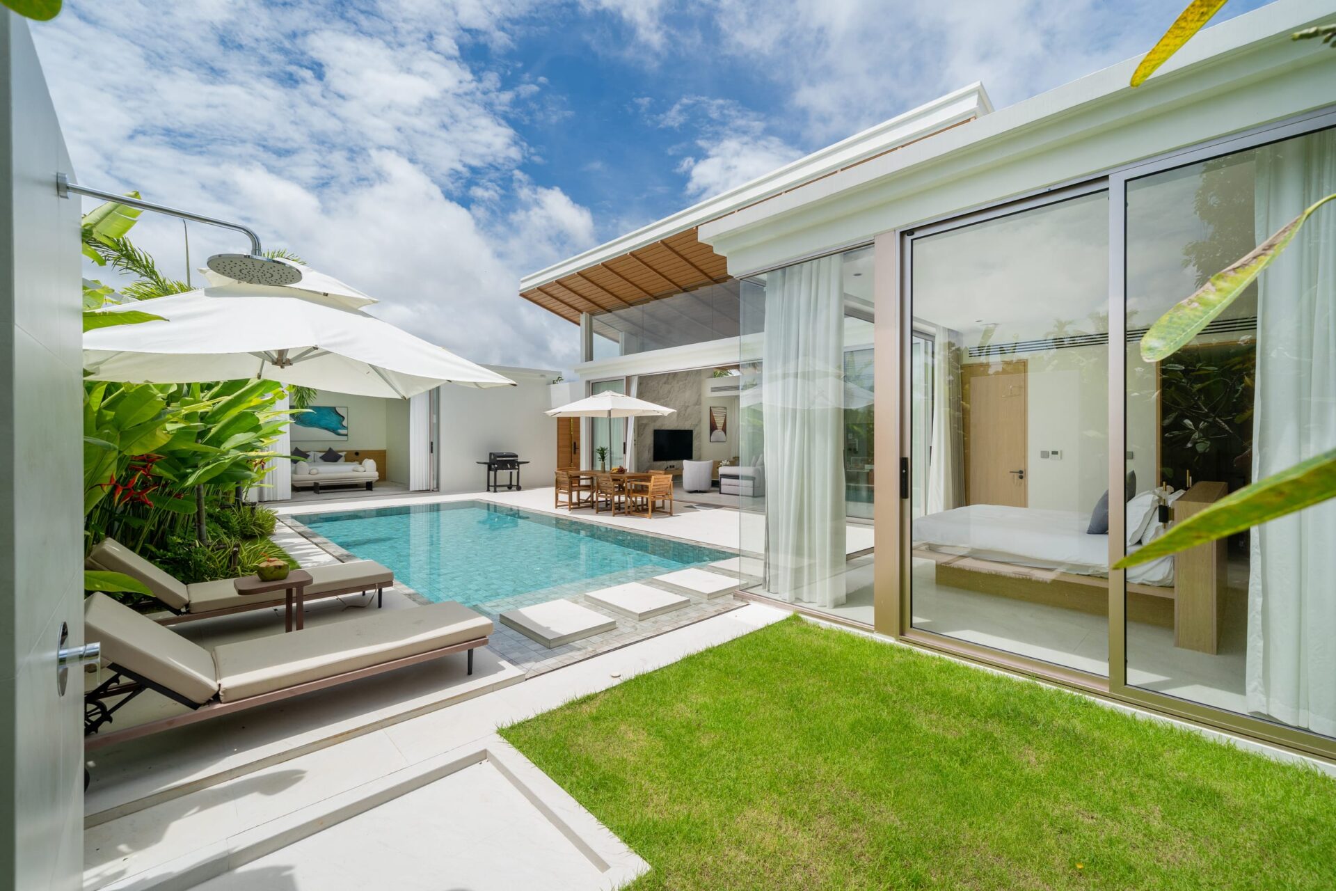 Modern villa backyard with a rectangular pool, white loungers, and umbrellas beside a glass-walled living area