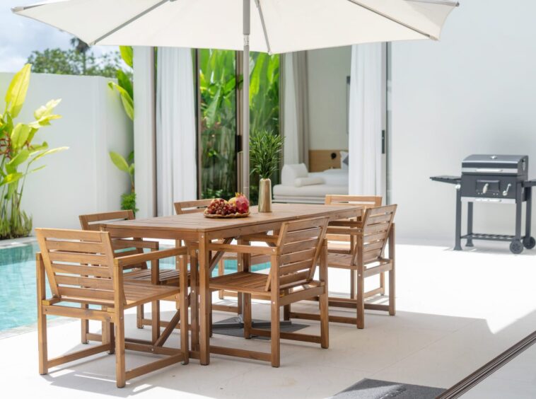 Outdoor dining area by a pool with a wooden table and chairs under a large white umbrella, a fruit platter in the center and a grill in the background.