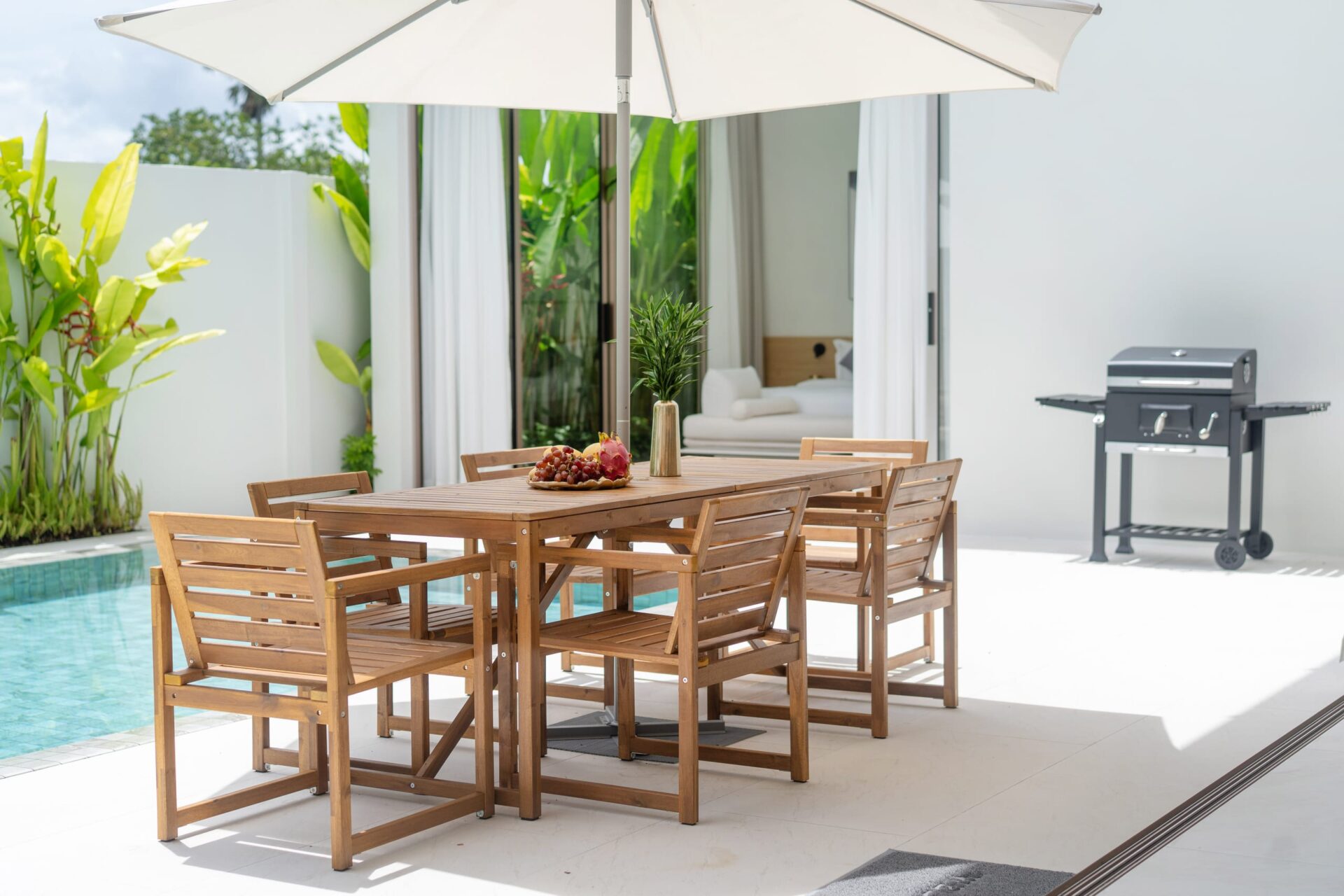 Outdoor dining area by a pool with a wooden table and chairs under a large white umbrella, a fruit platter in the center and a grill in the background.
