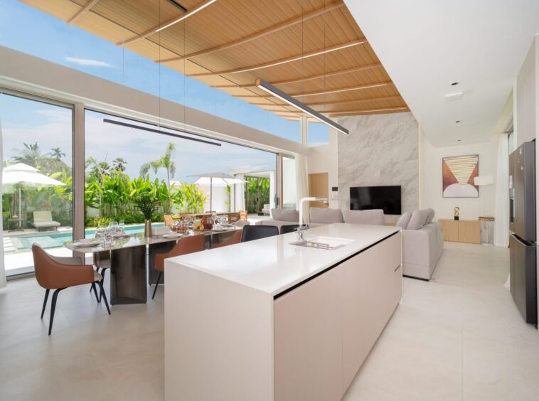 Open-plan kitchen and living area with an island, dining table, and glass doors opening to a pool and tropical garden. (Informative)