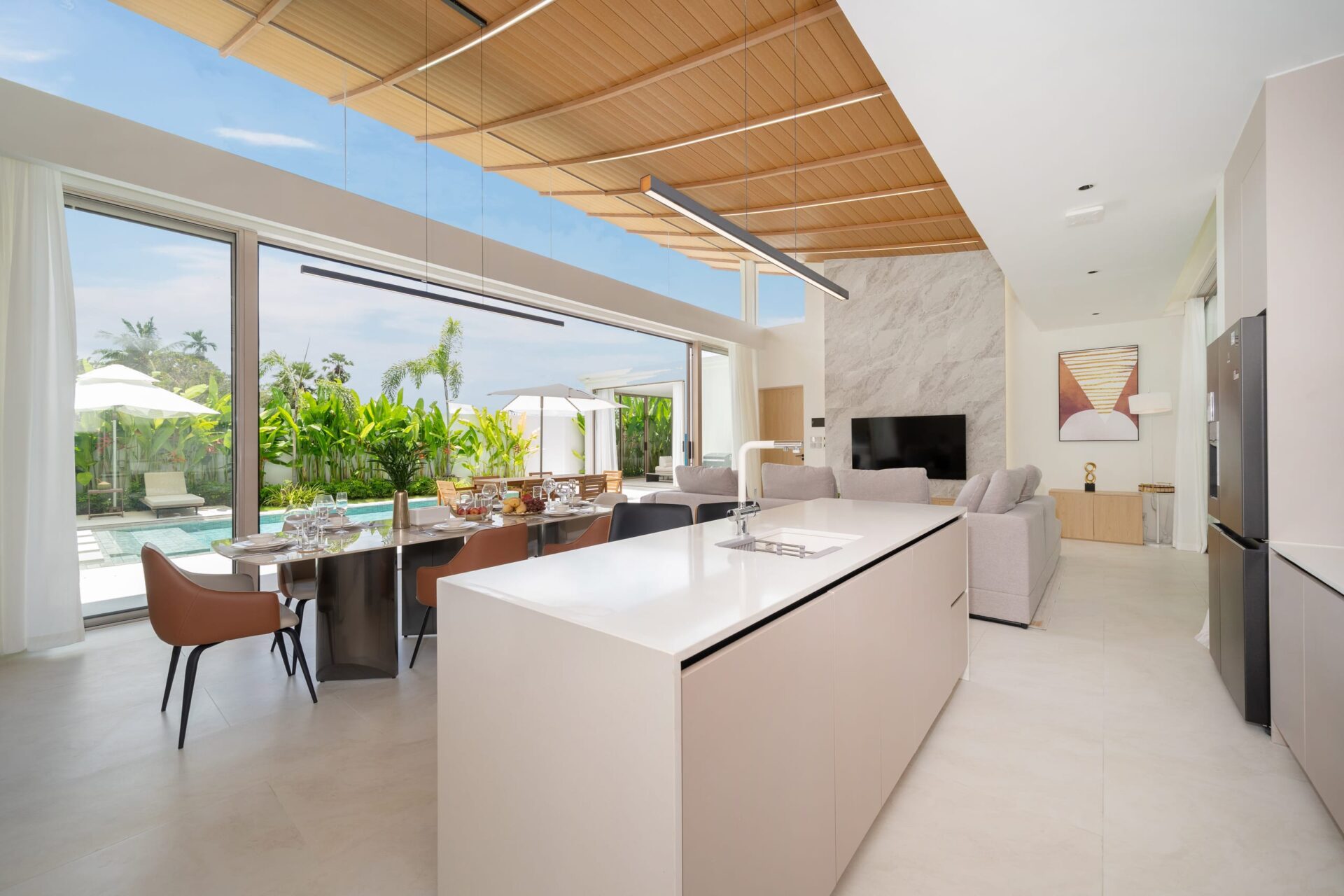 Open-plan kitchen and living area with an island, dining table, and glass doors opening to a pool and tropical garden. (Informative)