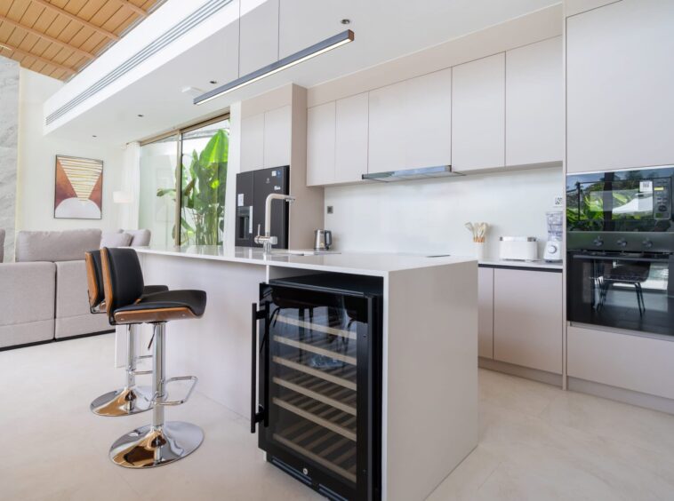 Modern white kitchen with an island, black-framed wine cooler, and bar stools, adjacent living area with a gray sofa and artwork.