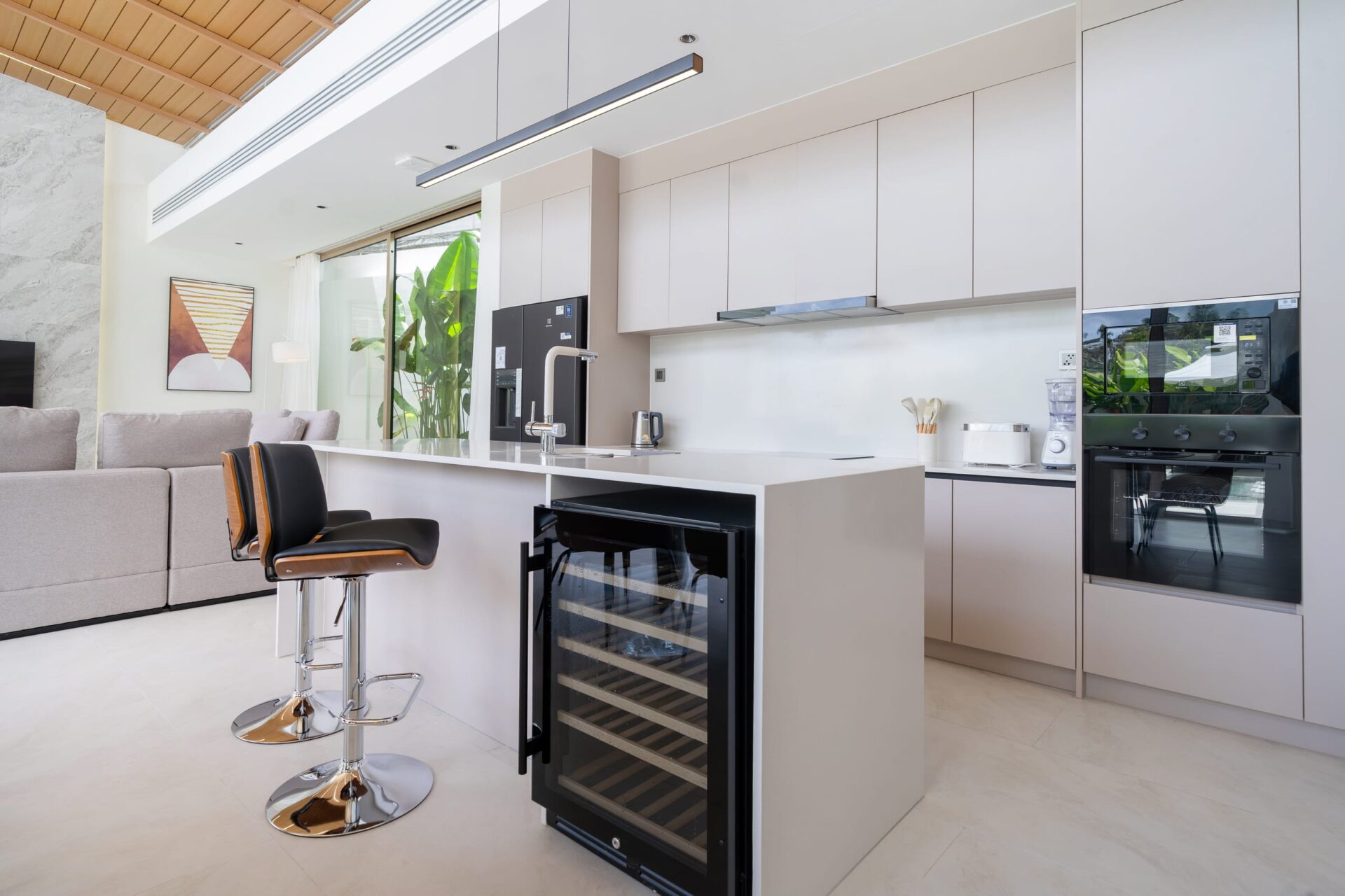 Modern white kitchen with an island, black-framed wine cooler, and bar stools, adjacent living area with a gray sofa and artwork.
