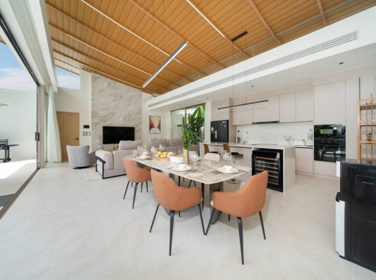 Open-plan kitchen and dining room with a warm wooden slat ceiling, marble table set for eight, and white cabinetry.