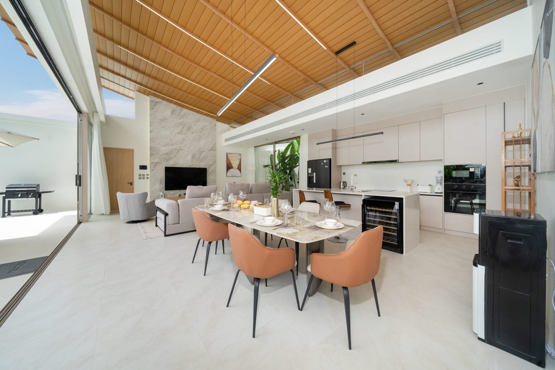 Open-plan kitchen and dining room with a warm wooden slat ceiling, marble table set for eight, and white cabinetry.