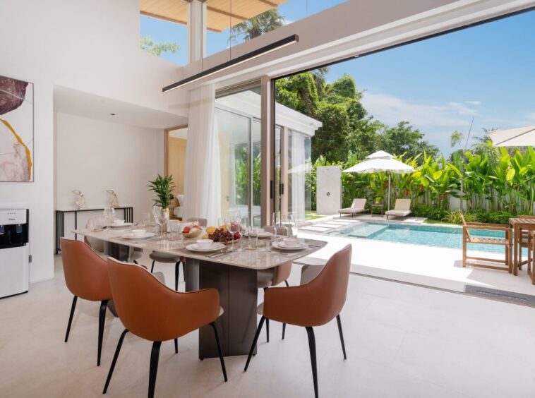 Open, sunlit dining area with a long table set for a meal, brown chairs, and glass sliding doors opening to a pool and tropical garden.